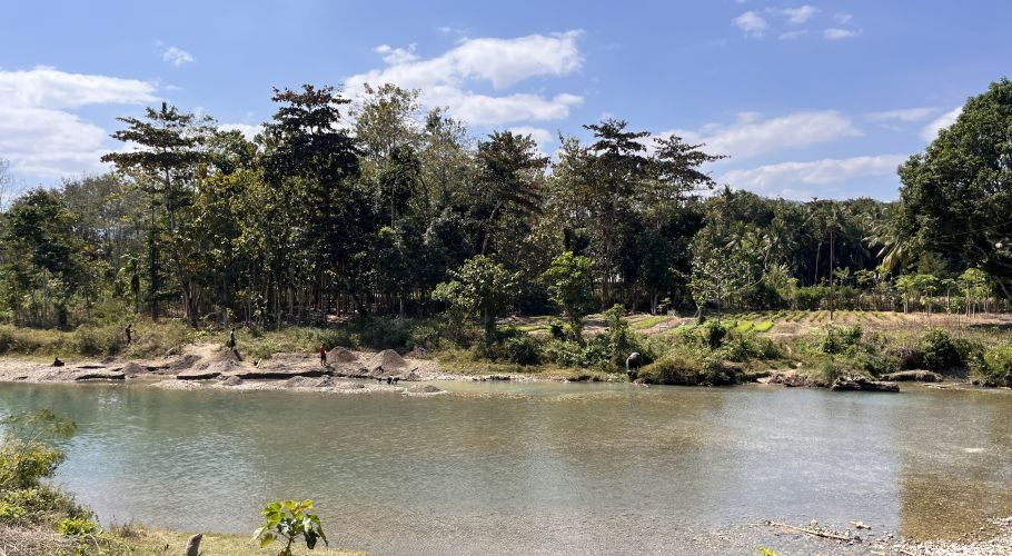 The wide Wanokaka River flows under a blue sky, with several people on its banks collecting stones amidst lush green trees and vegetation.