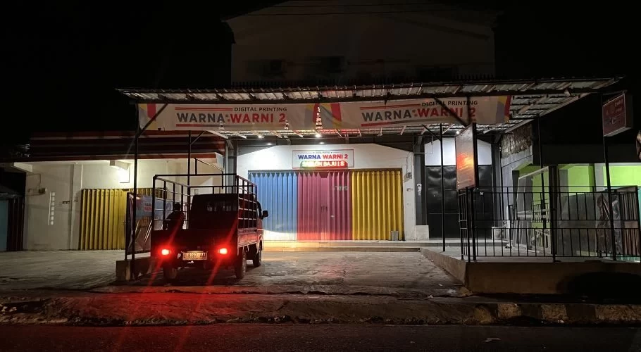 A dark pickup truck with red taillights is parked in front of the brightly lit "Warna Warni 2" printing shop at night, featuring colorful rolling doors.