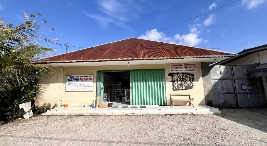 The Warna Warni Water Refill building in Loli, West Sumba, features light yellow walls, a rusty brown corrugated roof, and a partially open green accordion door under a clear blue sky.