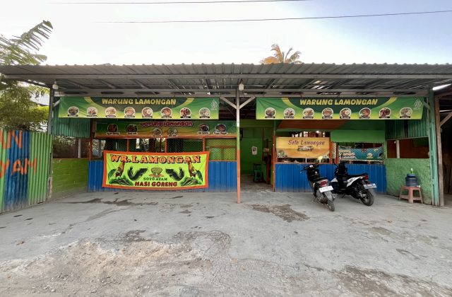 A green and blue street-side food stall, "Warung Lamongan," is visible under a metal roof with two motorbikes parked on the concrete ground.