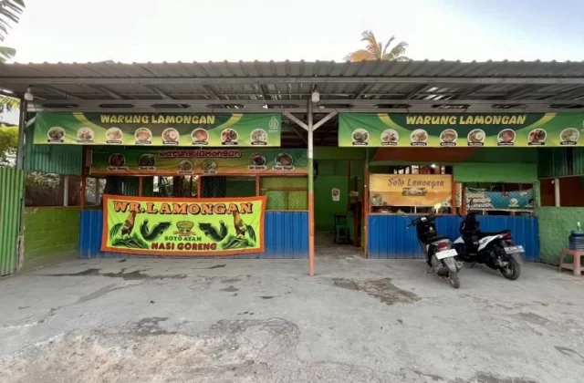 A green and blue street-side food stall, "Warung Lamongan," is visible under a metal roof with two motorbikes parked on the concrete ground.