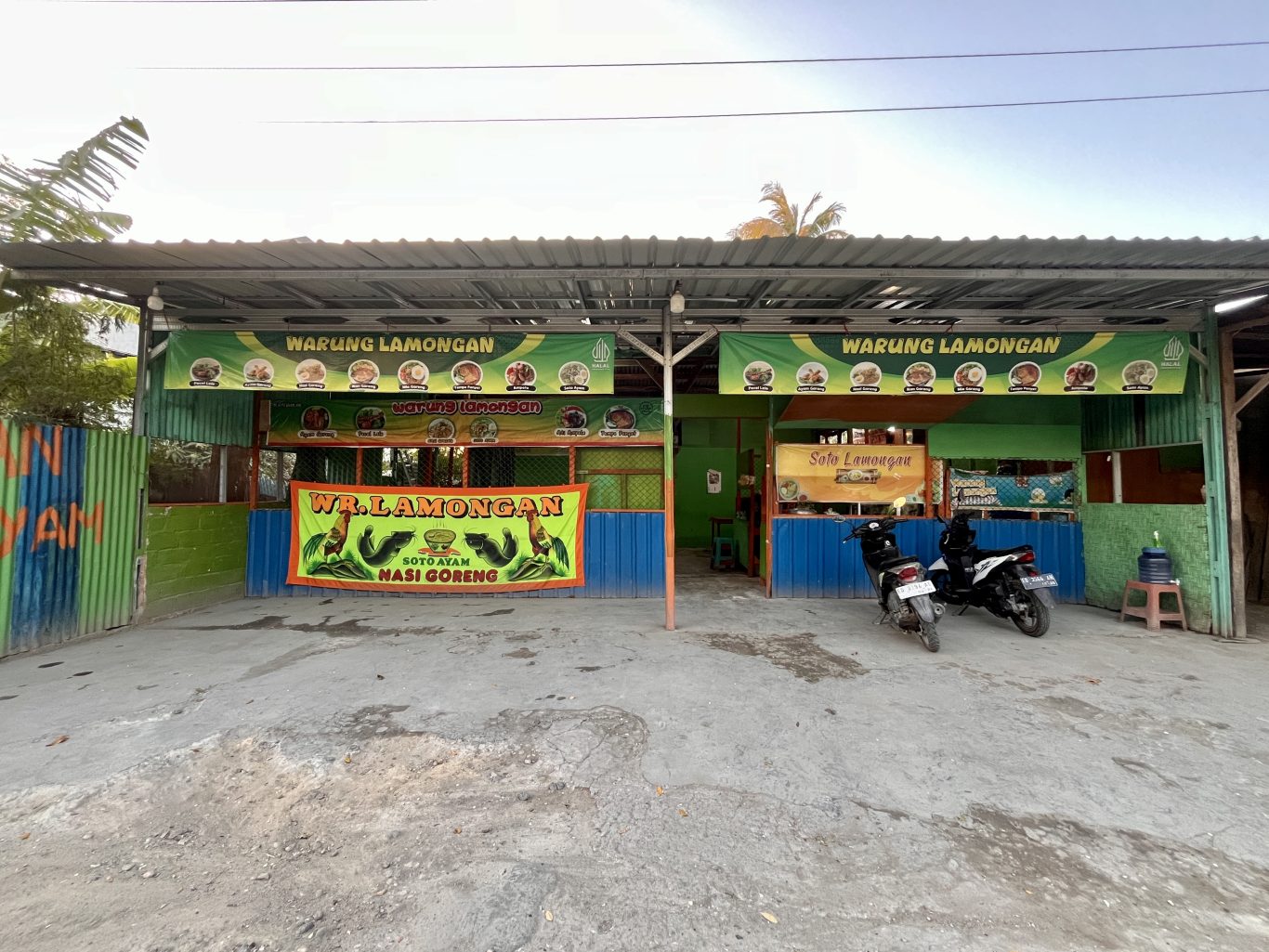 A green and blue street-side food stall, "Warung Lamongan," is visible under a metal roof with two motorbikes parked on the concrete ground.