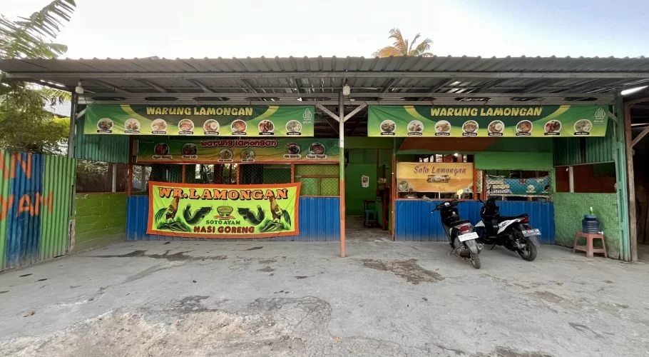 A green and blue street-side food stall, "Warung Lamongan," is visible under a metal roof with two motorbikes parked on the concrete ground.