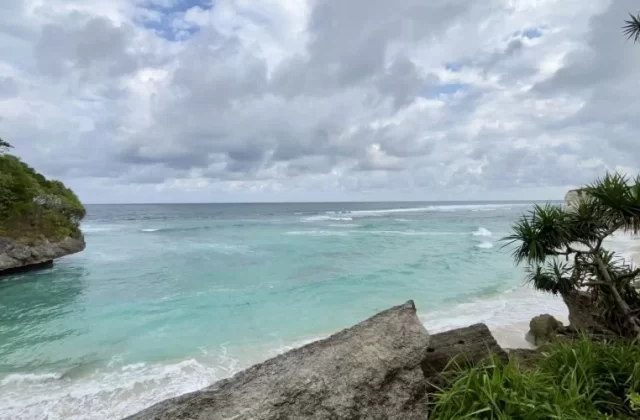 Turquoise ocean waves gently break on a white sand beach framed by green-topped rocky cliffs under a cloudy sky.