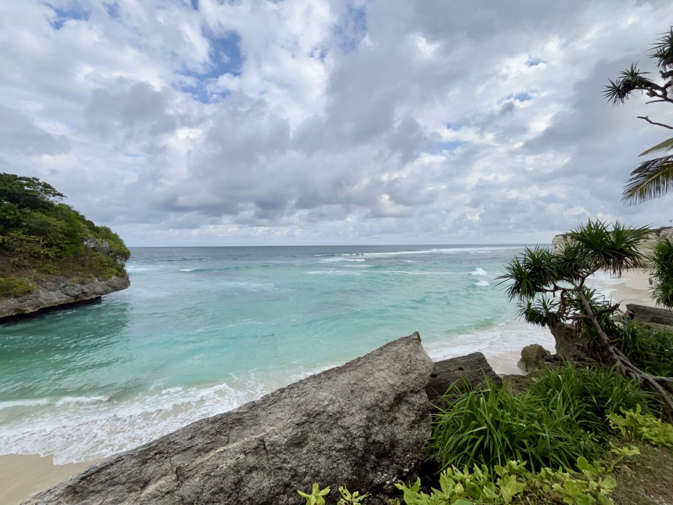 Turquoise ocean waves gently break on a white sand beach framed by green-topped rocky cliffs under a cloudy sky.