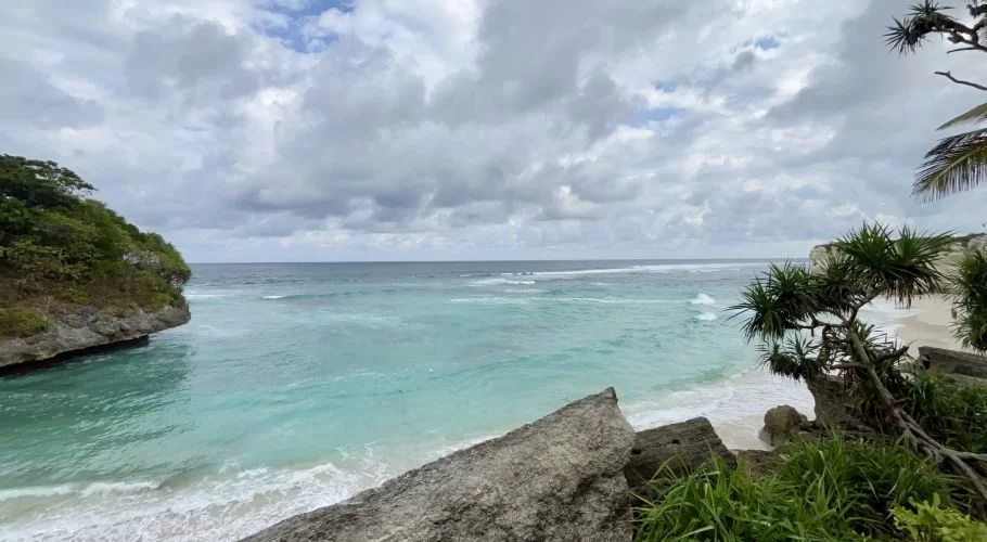 Turquoise ocean waves gently break on a white sand beach framed by green-topped rocky cliffs under a cloudy sky.