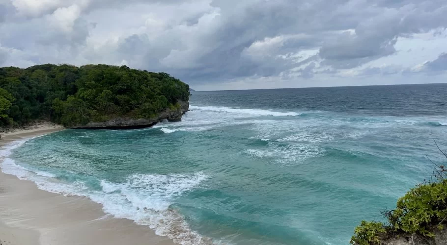 Turquoise ocean waves breaking on a white sand beach, bordered by a lush green, rocky headland under a cloudy sky.