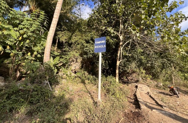 A blue sign for Kampung Wehola stands beside a winding cement path leading uphill through dense green foliage under a bright sky.
