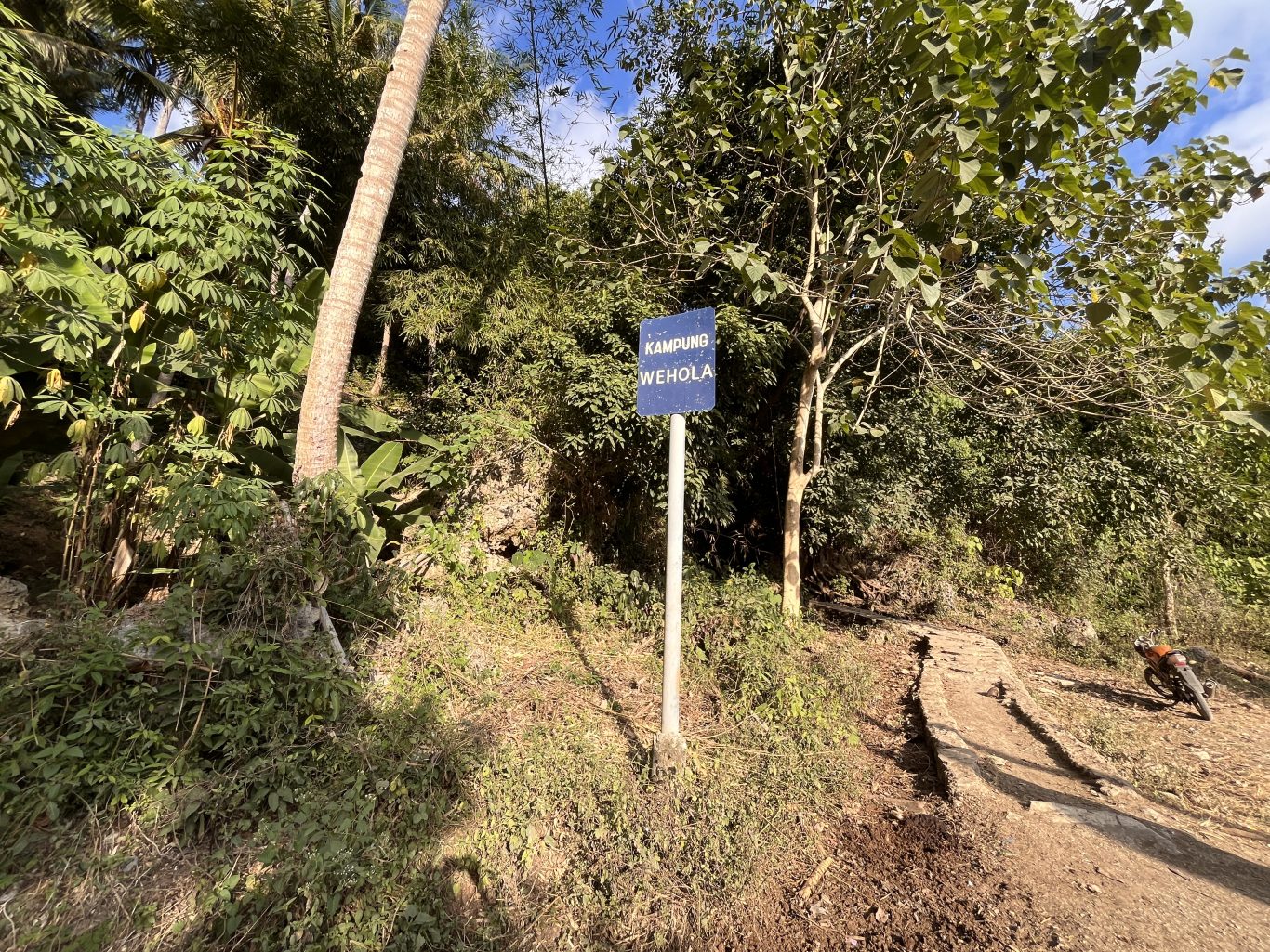 A blue sign for Kampung Wehola stands beside a winding cement path leading uphill through dense green foliage under a bright sky.