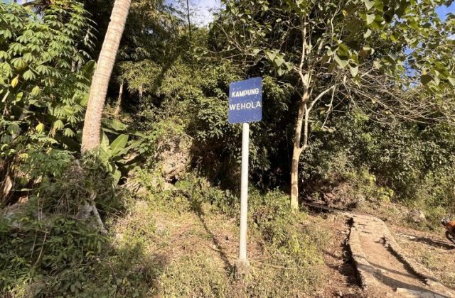 A blue sign for Kampung Wehola stands beside a winding cement path leading uphill through dense green foliage under a bright sky.