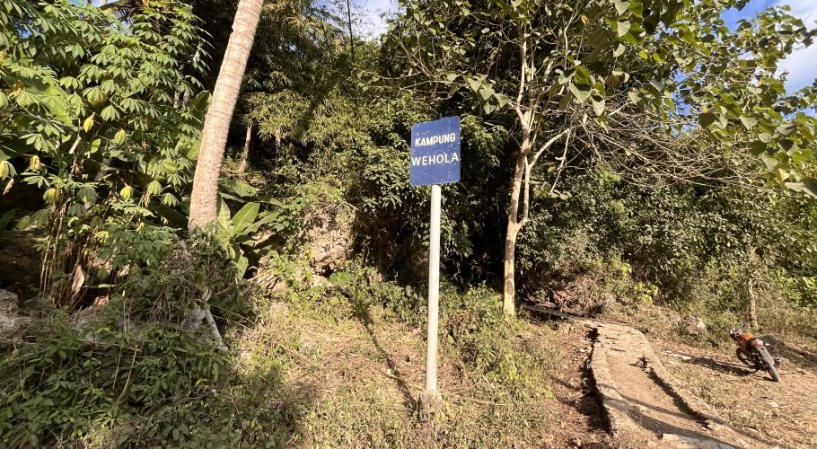 A blue sign for Kampung Wehola stands beside a winding cement path leading uphill through dense green foliage under a bright sky.