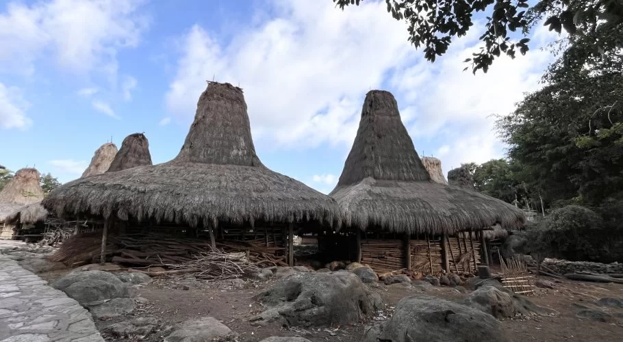 Traditional Sumba houses with tall, conical thatched roofs stand among large rocks and green trees under a partly cloudy blue sky.
