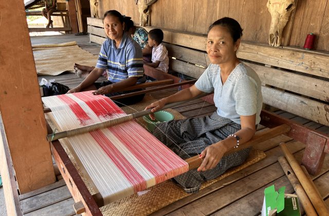 Two Indonesian women from the Wenei Weaving Group weave red and white patterned textiles on traditional looms inside a wooden house.