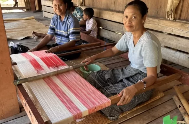 Two Indonesian women from the Wenei Weaving Group weave red and white patterned textiles on traditional looms inside a wooden house.