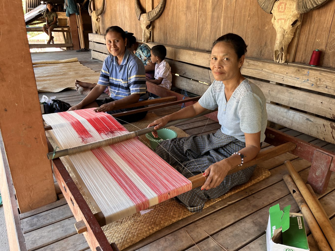 Two Indonesian women from the Wenei Weaving Group weave red and white patterned textiles on traditional looms inside a wooden house.