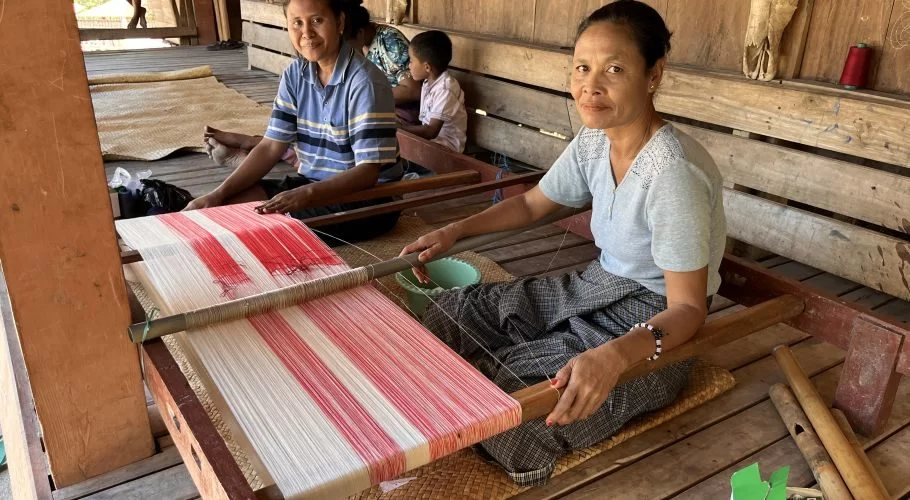 Two Indonesian women from the Wenei Weaving Group weave red and white patterned textiles on traditional looms inside a wooden house.