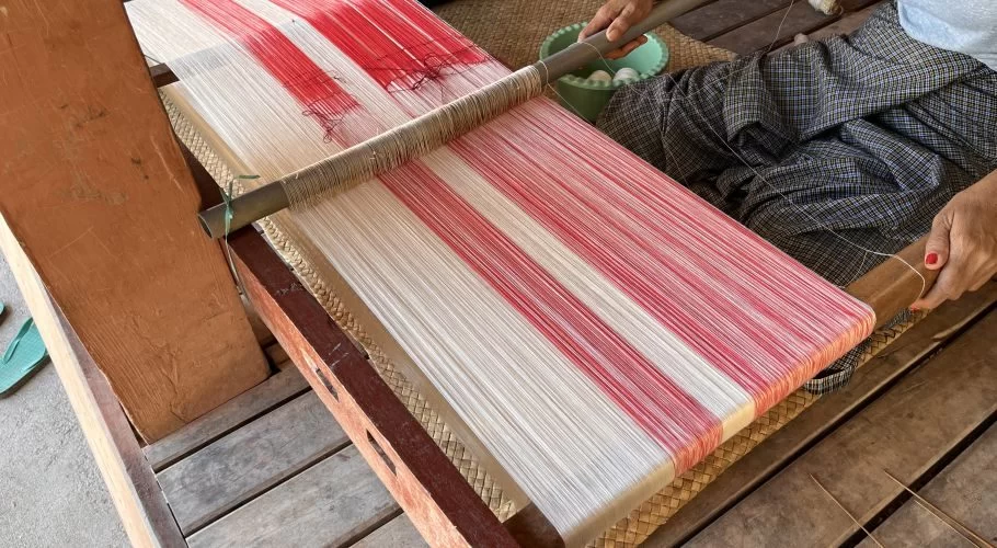 A person's hands weave red and white threads on a traditional wooden loom, sitting on a mat over a wooden floor.