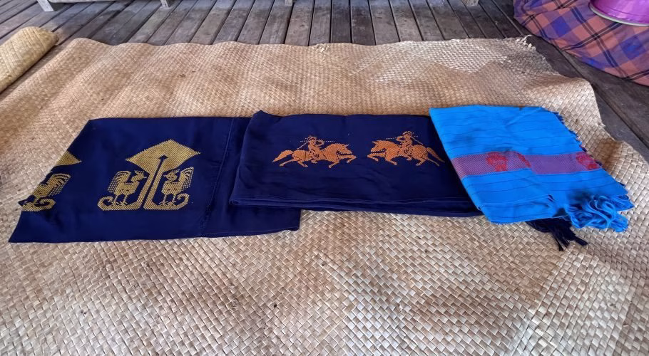 Three handcrafted textiles, dark blue with golden chicken and orange horse motifs, and a bright blue fringed piece, lie on a woven mat.