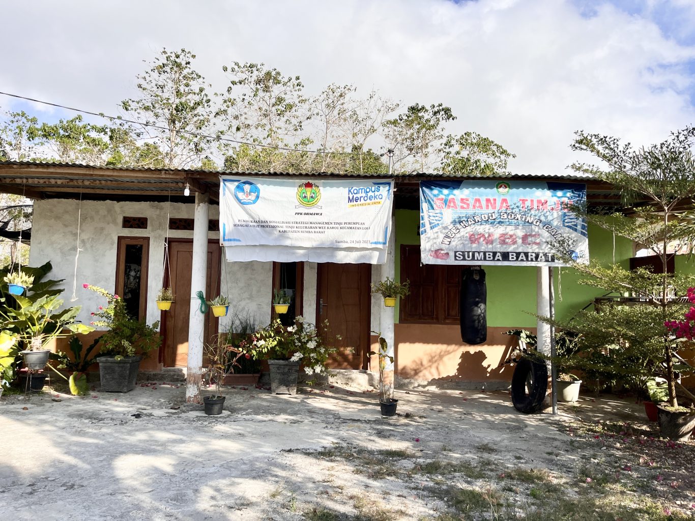 A modest, light-colored building with a corrugated roof displays two banners, one for Wee Karou Boxing Club, and a black punching bag hangs outside.