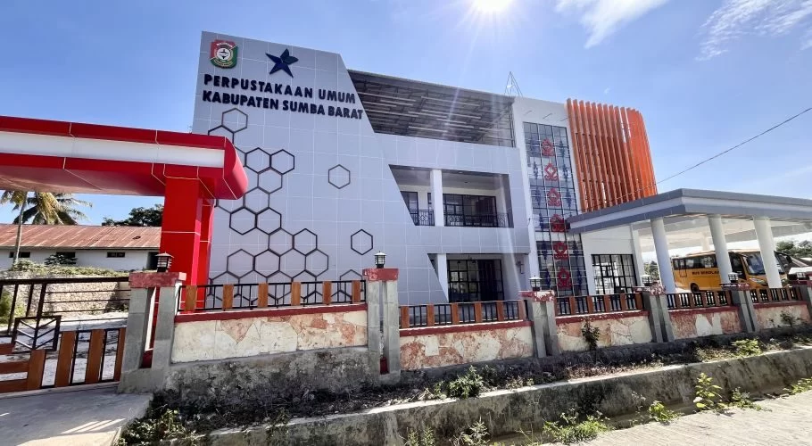 The modern, three-story West Sumba Public Library building features a white facade with a honeycomb pattern, red entrance, and orange vertical accents under a bright blue sky.