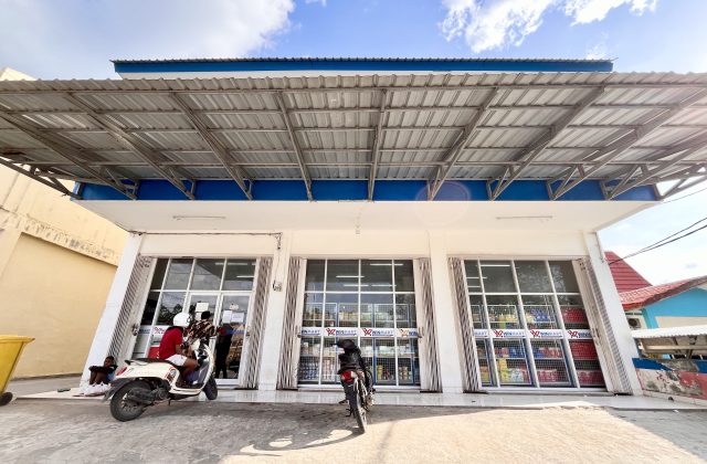 The white Winmart supermarket in Tambolaka, Sumba, features large glass windows, a blue trim, and a wide metal awning, with motorcycles parked outside.