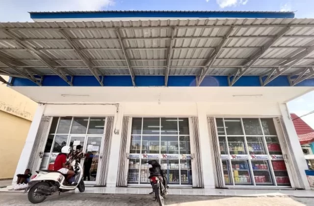 The white Winmart supermarket in Tambolaka, Sumba, features large glass windows, a blue trim, and a wide metal awning, with motorcycles parked outside.
