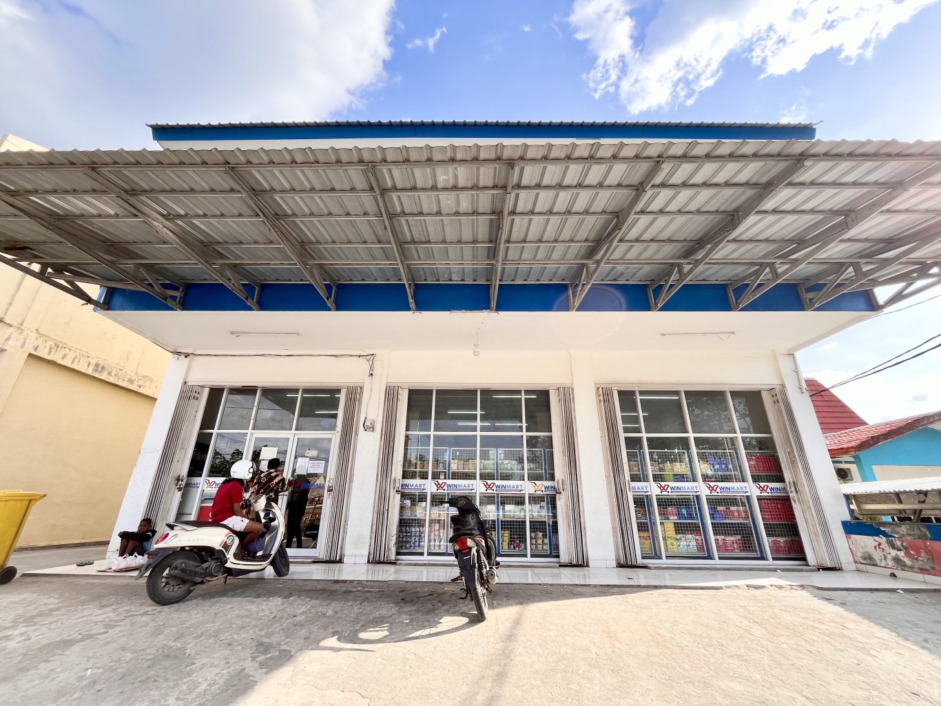 The white Winmart supermarket in Tambolaka, Sumba, features large glass windows, a blue trim, and a wide metal awning, with motorcycles parked outside.