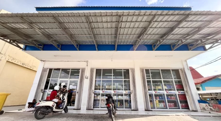 The white Winmart supermarket in Tambolaka, Sumba, features large glass windows, a blue trim, and a wide metal awning, with motorcycles parked outside.