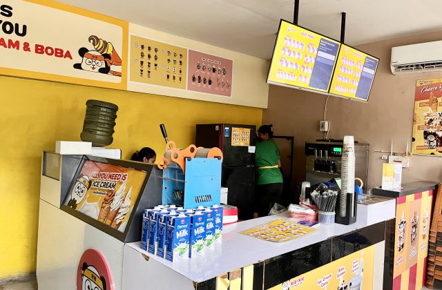Inside Xie-Xie Ice Cream & Boba, two staff members work behind a counter with ice cream machines, menu boards, and milk cartons against a vibrant yellow wall.