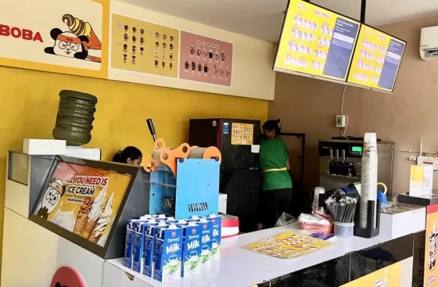 Inside Xie-Xie Ice Cream & Boba, two staff members work behind a counter with ice cream machines, menu boards, and milk cartons against a vibrant yellow wall.