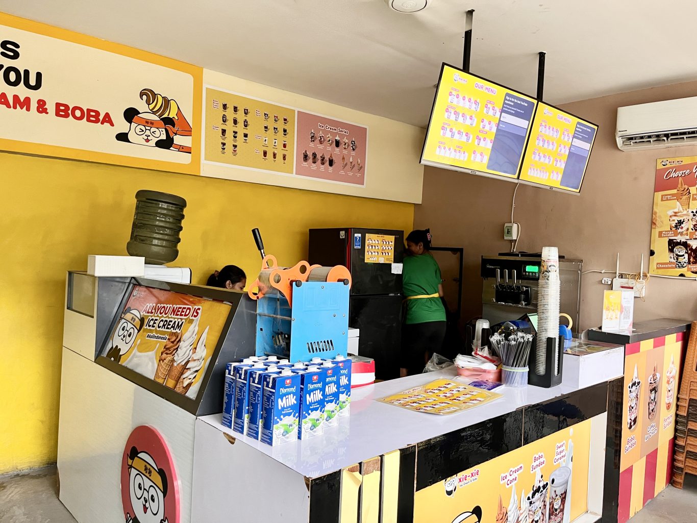 Inside Xie-Xie Ice Cream & Boba, two staff members work behind a counter with ice cream machines, menu boards, and milk cartons against a vibrant yellow wall.