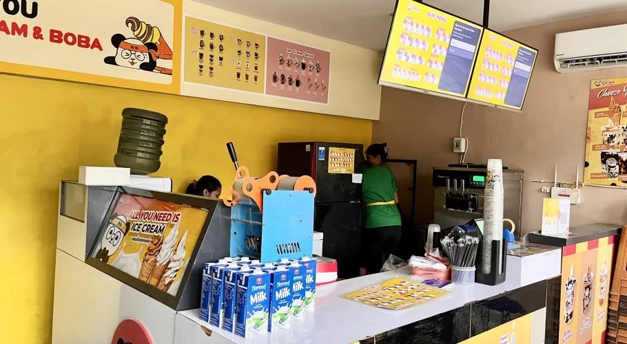 Inside Xie-Xie Ice Cream & Boba, two staff members work behind a counter with ice cream machines, menu boards, and milk cartons against a vibrant yellow wall.