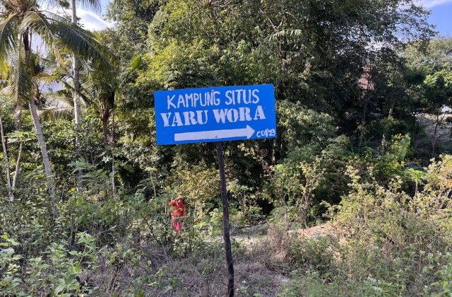 A blue sign with white text "KAMPUNG SITUS YARU WORA" and a right-pointing arrow stands amidst lush green tropical trees and foliage under a clear sky.