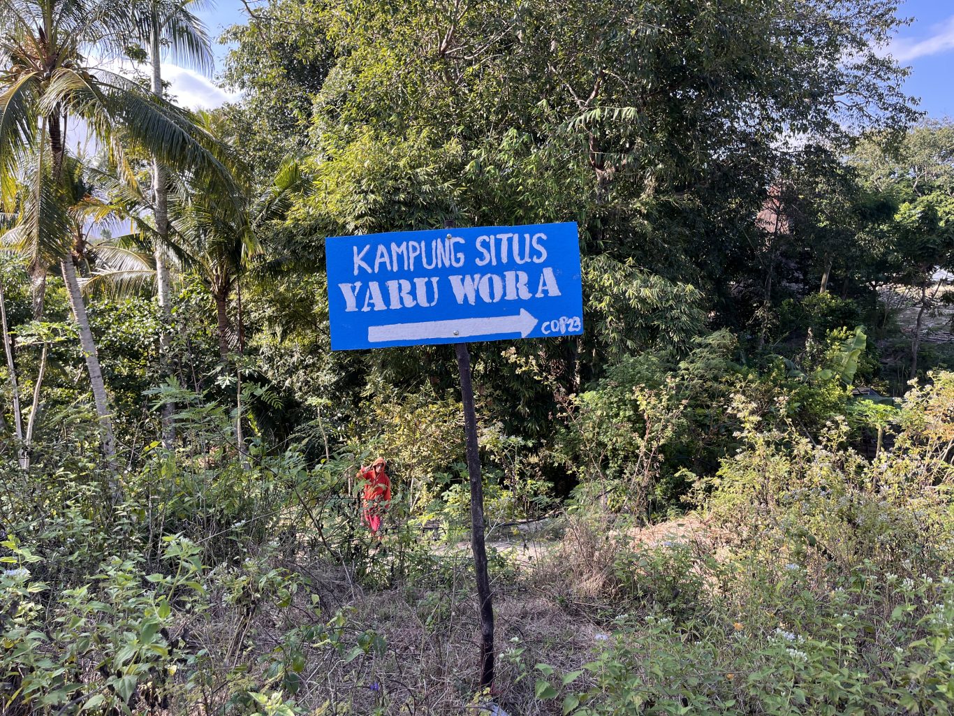A blue sign with white text "KAMPUNG SITUS YARU WORA" and a right-pointing arrow stands amidst lush green tropical trees and foliage under a clear sky.