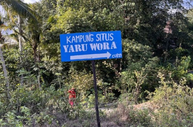 A blue sign with white text "KAMPUNG SITUS YARU WORA" and a right-pointing arrow stands amidst lush green tropical trees and foliage under a clear sky.