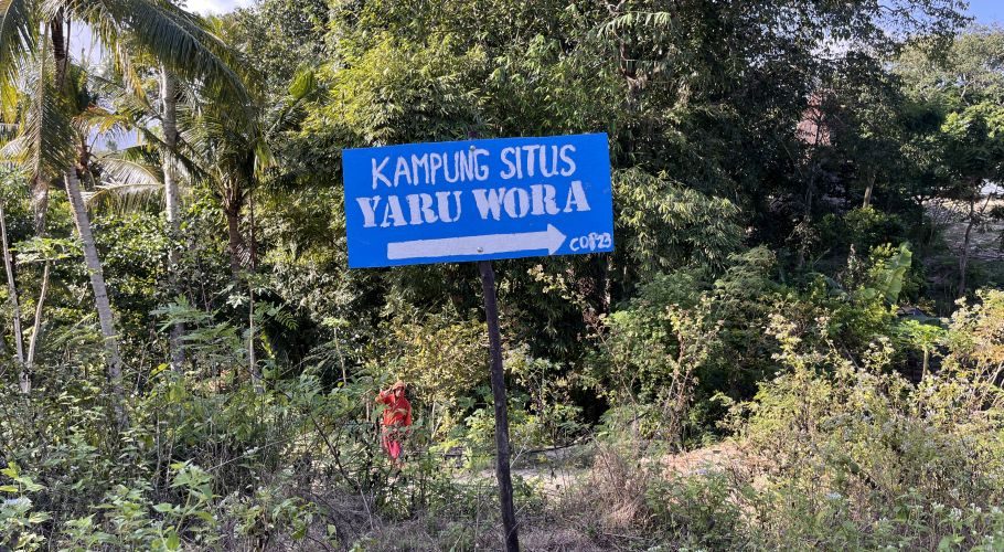 A blue sign with white text "KAMPUNG SITUS YARU WORA" and a right-pointing arrow stands amidst lush green tropical trees and foliage under a clear sky.