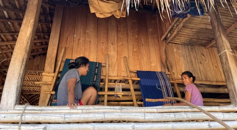 Two women are inside a traditional Sumbanese house, with one demonstrating weaving techniques on a loom with blue and green textiles.