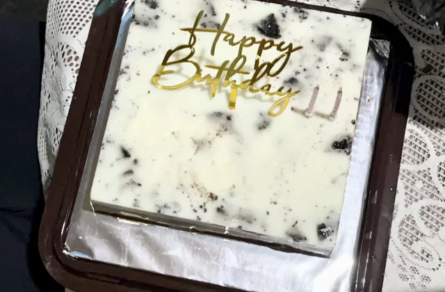 A square birthday cake with a white and dark cookie crumb topping, a gold "Happy Birthday" topper, and two unlit candles, presented in a brown tray on a lace tablecloth.