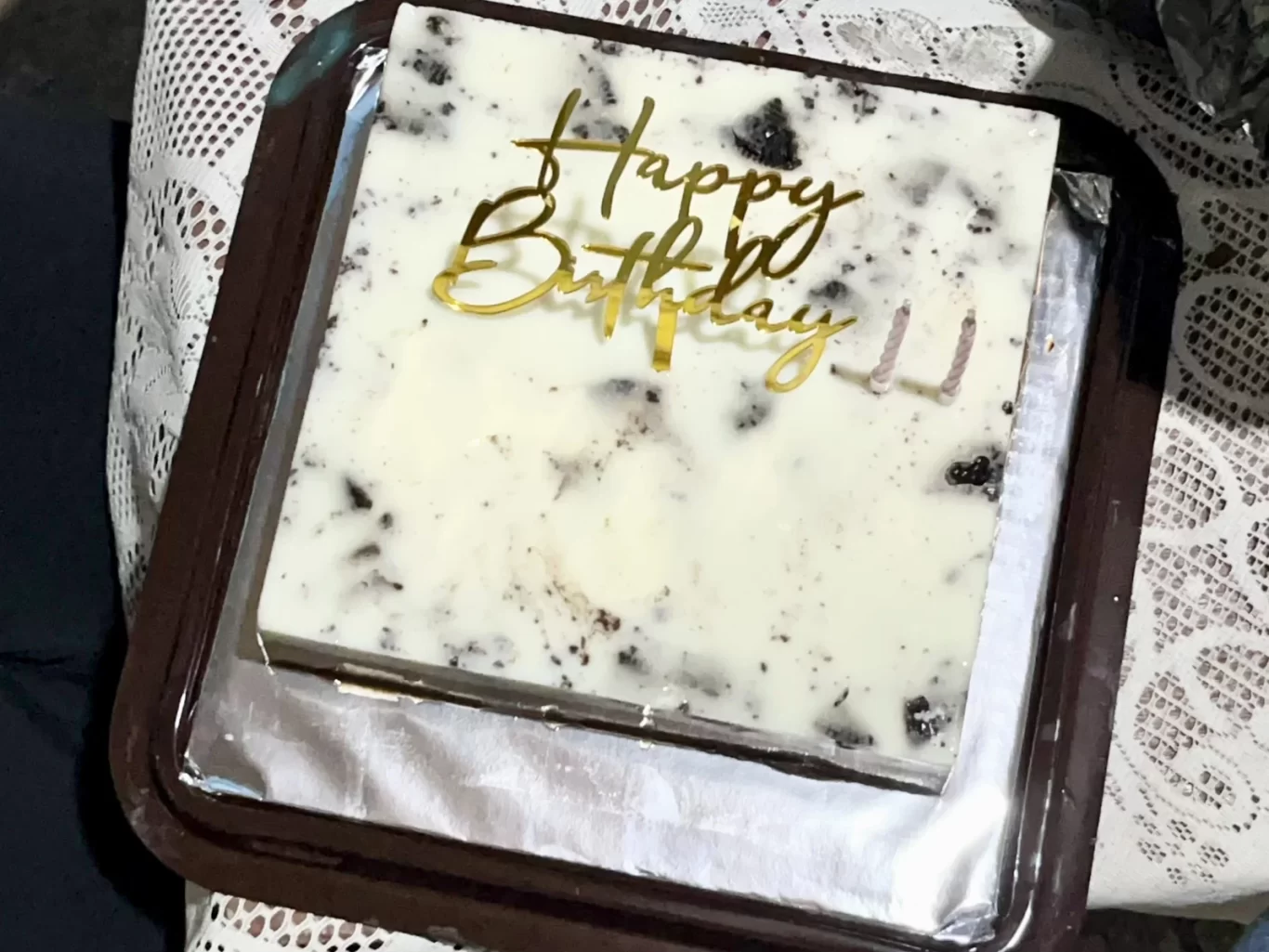 A square birthday cake with a white and dark cookie crumb topping, a gold "Happy Birthday" topper, and two unlit candles, presented in a brown tray on a lace tablecloth.