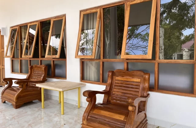 Two ornate wooden chairs and a small table sit on a tiled patio in front of a white wall with open wooden-framed windows reflecting trees.