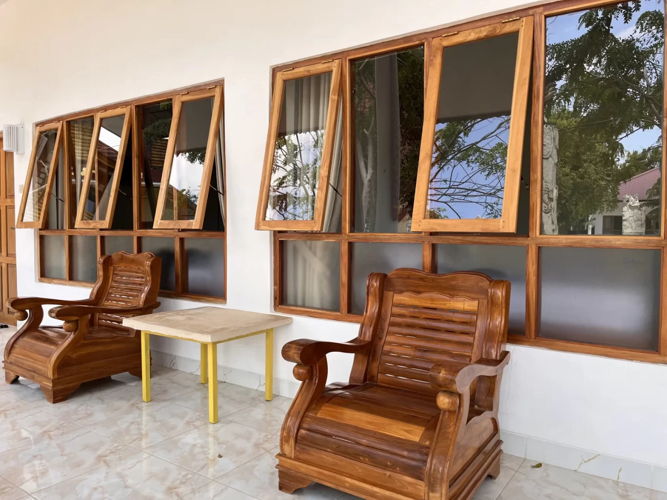 Two ornate wooden chairs and a small table sit on a tiled patio in front of a white wall with open wooden-framed windows reflecting trees.