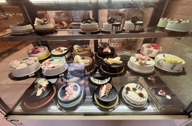 A refrigerated display case at King Bakery Tambolaka showcases numerous round cakes with colorful frostings and decorations.