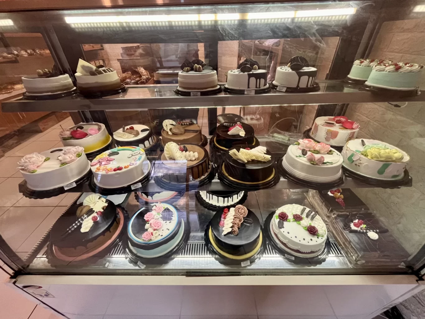 A refrigerated display case at King Bakery Tambolaka showcases numerous round cakes with colorful frostings and decorations.