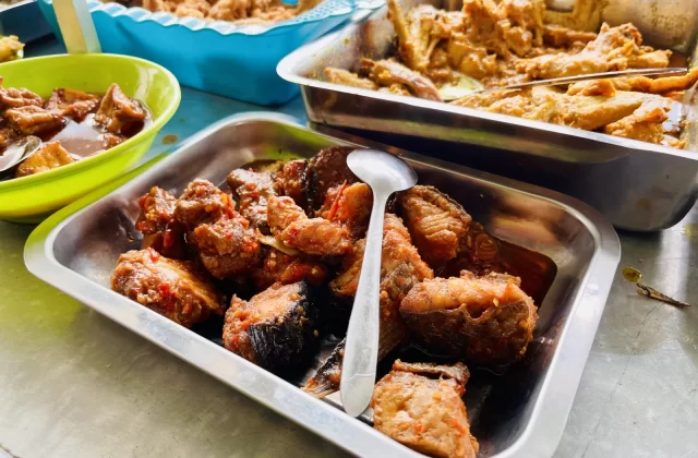 Multiple metal trays and a green bowl display various Indonesian dishes, including saucy fish, chicken, and fried items, on a light counter.