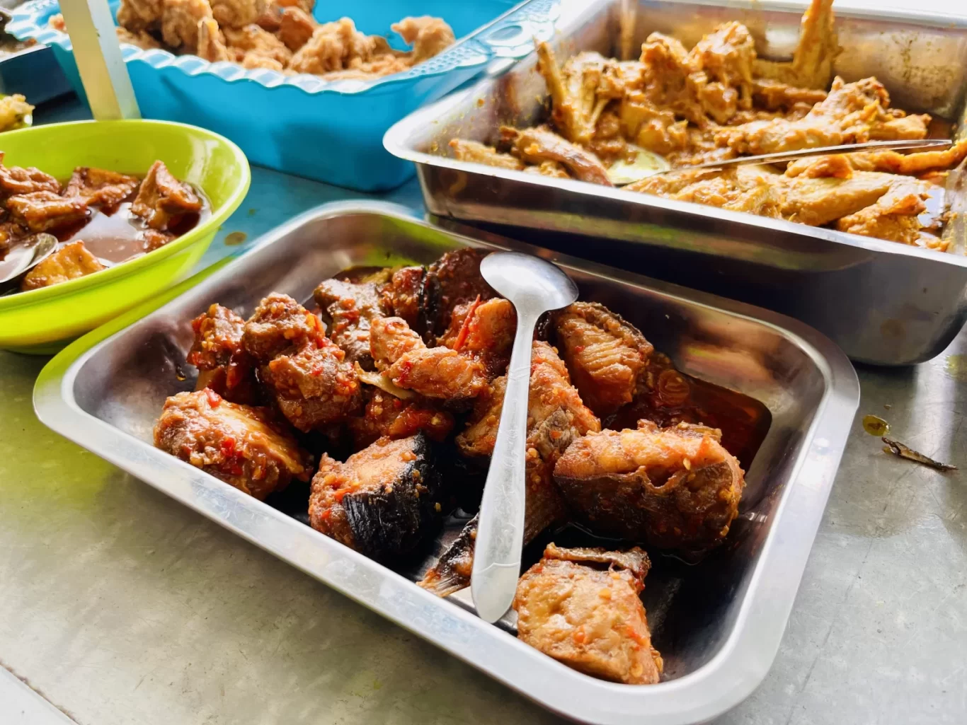Multiple metal trays and a green bowl display various Indonesian dishes, including saucy fish, chicken, and fried items, on a light counter.