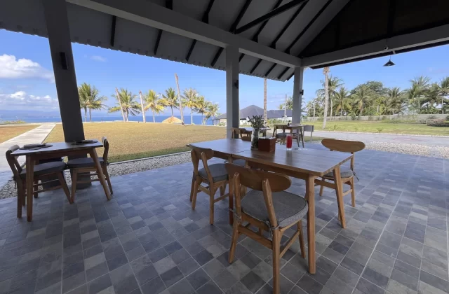 A restaurant patio with wooden tables and chairs features a tiled floor and overlooks a green lawn, palm trees, and a blue ocean under a clear sky.