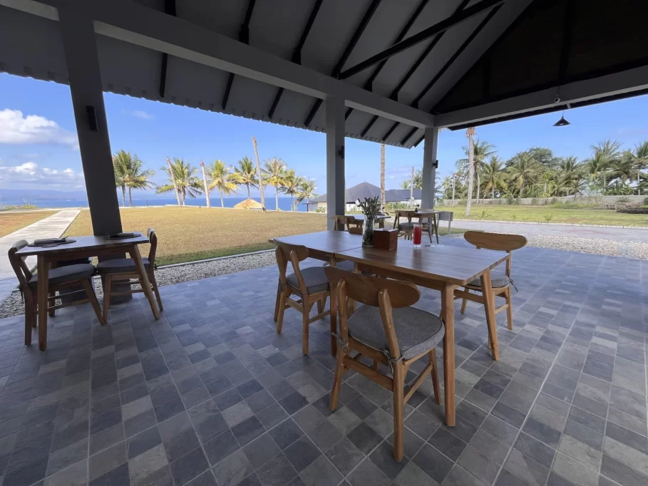 A restaurant patio with wooden tables and chairs features a tiled floor and overlooks a green lawn, palm trees, and a blue ocean under a clear sky.