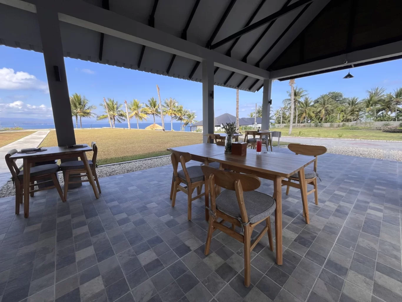 A restaurant patio with wooden tables and chairs features a tiled floor and overlooks a green lawn, palm trees, and a blue ocean under a clear sky.