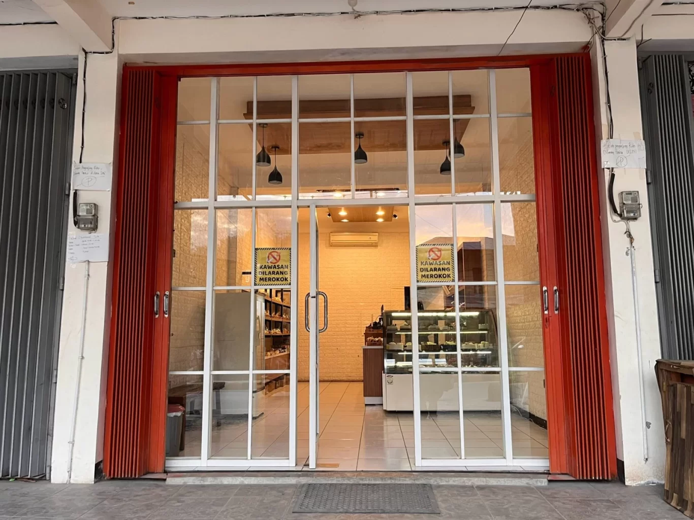 The storefront of King Bakery Tambolaka features large glass doors with white frames, flanked by partially open red accordion security gates, revealing warm interior lighting and pastry displays.