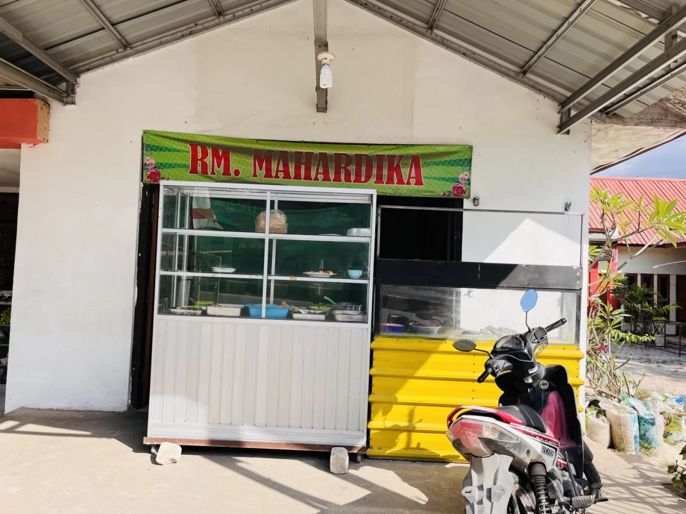 The white exterior of Mahardika Restaurant in Sumba features a green banner, a glass food display case, and a yellow panel, with a scooter parked in front.